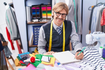 Téléchargez les photos : Jeune homme blond tailleur souriant dessin confiant sur carnet à l'usine de vêtements - en image libre de droit