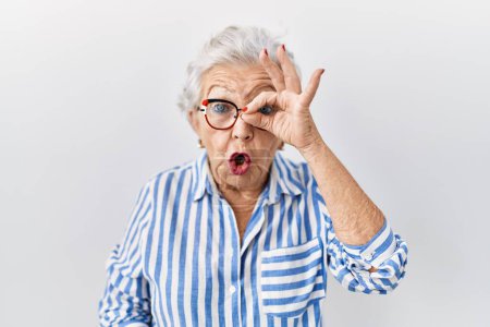 Téléchargez les photos : Femme âgée aux cheveux gris debout sur fond blanc faisant un geste correct choqué par le visage surpris, les yeux regardant à travers les doigts. expression incrédule. - en image libre de droit