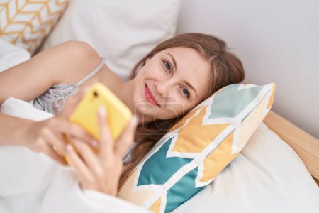 Téléchargez les photos : Jeune femme caucasienne utilisant smartphone couché sur le lit dans la chambre - en image libre de droit