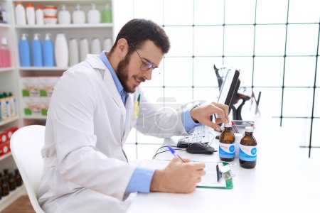 Foto de Joven farmacéutico hispano sosteniendo frasco de medicación escribiendo en documento en farmacia - Imagen libre de derechos