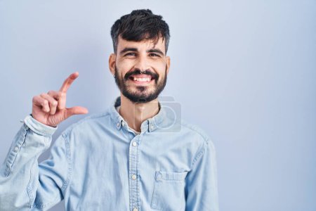 Téléchargez les photos : Jeune homme hispanique avec la barbe debout sur fond bleu geste souriant et confiant avec la main faisant signe de petite taille avec les doigts regardant et la caméra. concept de mesure. - en image libre de droit