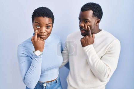 Téléchargez les photos : Jeune couple afro-américain debout sur fond bleu pointant vers l'œil vous regardant geste, expression suspecte - en image libre de droit