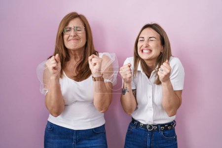 Foto de Madre e hija hispanas vistiendo una camiseta blanca casual sobre fondo rosa emocionadas por el éxito con los brazos levantados y los ojos cerrados celebrando la victoria sonriendo. concepto ganador. - Imagen libre de derechos