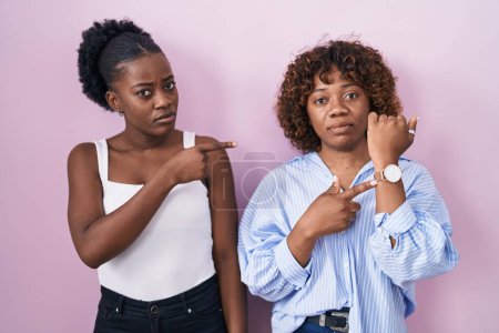Téléchargez les photos : Deux femmes africaines debout sur fond rose pressé pointant vers regarder le temps, l'impatience, regardant la caméra avec une expression détendue - en image libre de droit