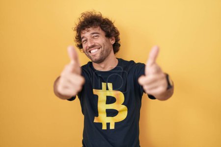 Téléchargez les photos : Jeune homme hispanique portant Bitcoin t-shirt pointant les doigts vers l'appareil photo avec visage heureux et drôle. bonne énergie et vibes. - en image libre de droit
