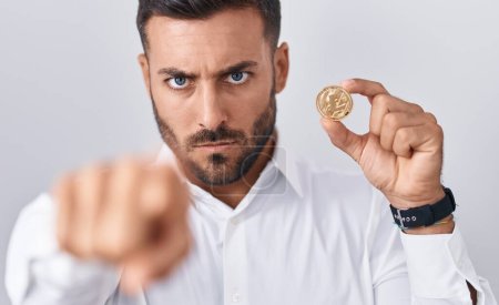 Téléchargez les photos : Bel homme hispanique tenant litecoin pièce crypto-monnaie pointant du doigt à la caméra et à vous, geste confiant regardant sérieux - en image libre de droit