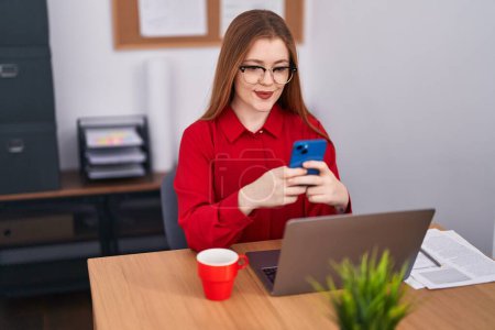 Téléchargez les photos : Jeune rousse femme d'affaires utilisant un ordinateur portable et un smartphone au bureau - en image libre de droit