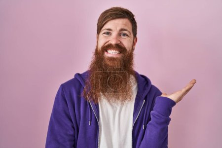 Téléchargez les photos : Homme caucasien avec longue barbe debout sur fond rose souriant gai présentation et pointant avec la paume de la main en regardant la caméra. - en image libre de droit