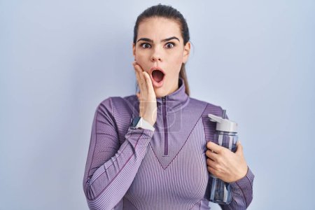 Photo for Beautiful woman wearing sportswear holding water bottle afraid and shocked, surprise and amazed expression with hands on face - Royalty Free Image