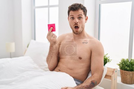 Foto de Joven hombre hispano sosteniendo condón en la cama asustado y sorprendido con la boca abierta para sorpresa, cara de incredulidad - Imagen libre de derechos