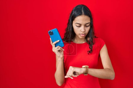 Foto de Mujer brasileña joven usando teléfono inteligente sobre fondo rojo comprobar la hora en reloj de pulsera, relajado y seguro - Imagen libre de derechos