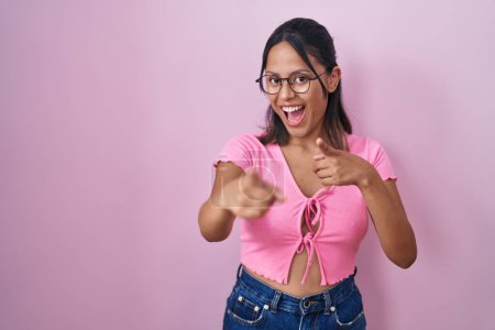 Foto de Mujer joven hispana de pie sobre fondo rosa con gafas que señalan con los dedos a la cámara con la cara feliz y divertida. buena energía y vibraciones. - Imagen libre de derechos