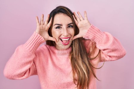 Foto de Joven mujer hispana de pie sobre fondo rosa sonriente alegre jugando a echar un vistazo a un boo con las manos mostrando la cara. sorprendido y salido - Imagen libre de derechos