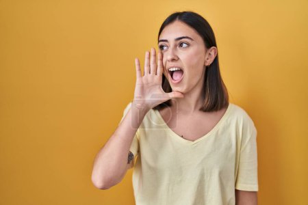 Foto de Chica hispana con camiseta casual sobre fondo amarillo gritando y gritando fuerte a un lado con la mano en la boca. concepto de comunicación. - Imagen libre de derechos