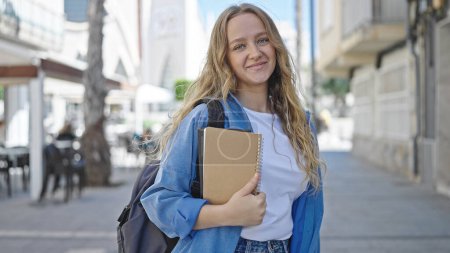 Téléchargez les photos : Jeune étudiante blonde souriante confiante tenant des livres dans la rue - en image libre de droit