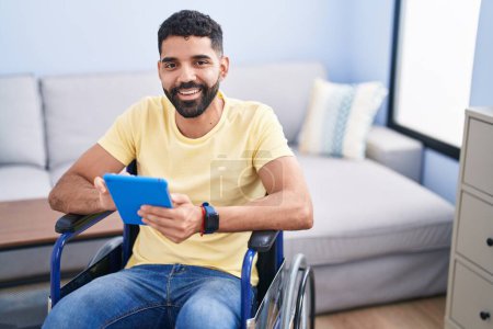 Téléchargez les photos : Young arab man using touchpad sitting on wheelchair at home - en image libre de droit