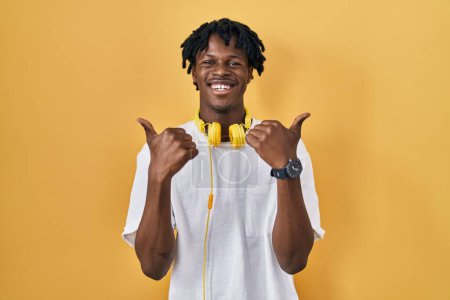 Foto de Hombre africano joven con rastas de pie sobre el signo de éxito de fondo amarillo haciendo gesto positivo con la mano, pulgares hacia arriba sonriendo y feliz. expresión alegre y gesto ganador. - Imagen libre de derechos