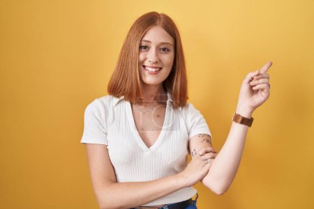 Photo for Young redhead woman standing over yellow background with a big smile on face, pointing with hand finger to the side looking at the camera. - Royalty Free Image
