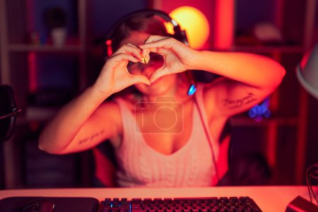 Photo for Young blonde woman playing video games wearing headphones smiling in love doing heart symbol shape with hands. romantic concept. - Royalty Free Image
