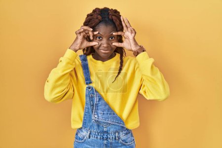 Téléchargez les photos : Femme africaine debout sur fond jaune essayant d'ouvrir les yeux avec les doigts, somnolent et fatigué pour la fatigue matinale - en image libre de droit