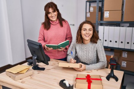 Téléchargez les photos : Two women ecommerce business workers writing on package reading book at office - en image libre de droit