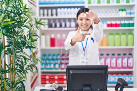 Téléchargez les photos : Jeune femme hispanique travaillant à la pharmacie pharmacie souriant faisant cadre avec les mains et les doigts avec le visage heureux. concept de créativité et photographie. - en image libre de droit