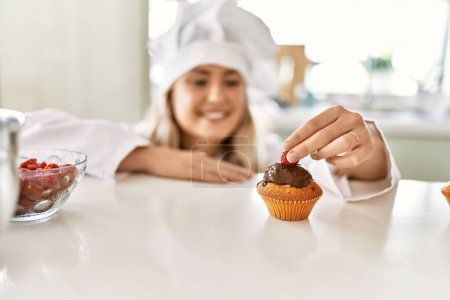 Photo for Young woman wearing cook uniform putting raspberry on cupcake at kitchen - Royalty Free Image