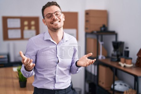 Foto de Joven hombre hispano en la oficina sonriendo alegre con los brazos abiertos como bienvenida amistosa, saludos positivos y confiados - Imagen libre de derechos