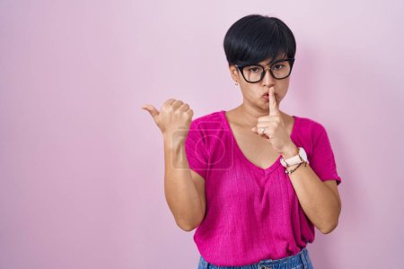 Foto de Mujer asiática joven con el pelo corto de pie sobre fondo rosa pidiendo estar tranquilo con el dedo en los labios señalando con la mano a un lado. silencio y concepto secreto. - Imagen libre de derechos