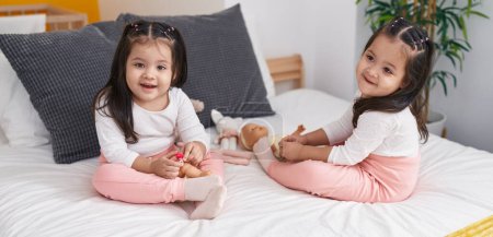 Foto de Adorables chicas gemelas jugando con la muñeca sentada en la cama en el dormitorio - Imagen libre de derechos
