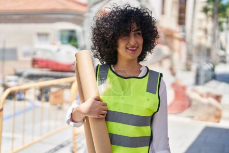 Foto de Joven arquitecta de Oriente Medio sonriendo confiada sosteniendo planos en la calle - Imagen libre de derechos