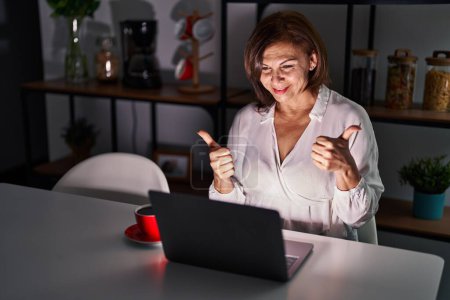Foto de Mujer hispana de mediana edad usando el ordenador portátil en casa por la noche signo de éxito haciendo gesto positivo con la mano, pulgares hacia arriba sonriendo y feliz. expresión alegre y gesto ganador. - Imagen libre de derechos