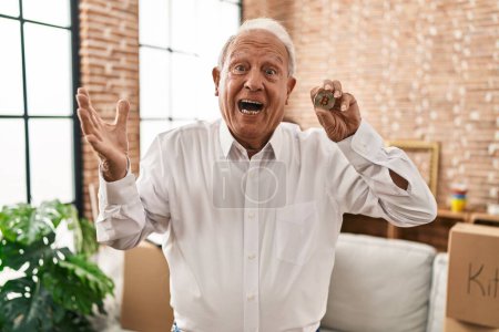 Foto de Hombre mayor con pelo gris sosteniendo moneda virtual bitcoin celebrando la victoria con sonrisa feliz y la expresión ganadora con las manos levantadas - Imagen libre de derechos