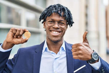 Foto de Joven hombre africano con rastas celebración de bitcoin moneda virtual sonriendo feliz y positivo, pulgar hacia arriba haciendo excelente y signo de aprobación - Imagen libre de derechos