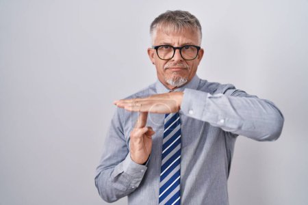 Foto de Hombre de negocios hispano con cabello gris usando gafas haciendo un gesto de tiempo fuera con las manos, rostro frustrado y serio - Imagen libre de derechos