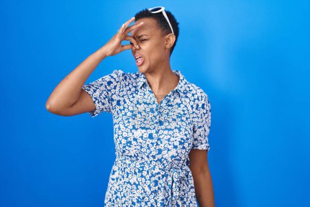 Téléchargez les photos : Femme afro-américaine debout sur fond bleu sentant quelque chose de puant et dégoûtant, odeur intolérable, retenant souffle avec les doigts sur le nez. mauvaise odeur - en image libre de droit
