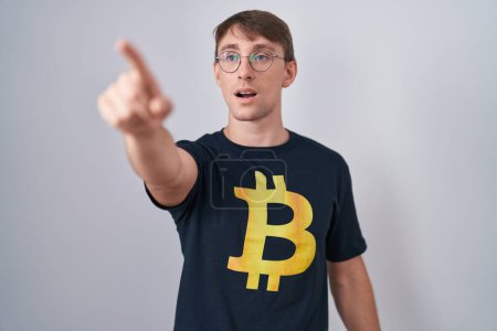 Foto de Hombre rubio caucásico con camiseta bitcoin apuntando con el dedo sorprendido por delante, boca abierta expresión asombrada, algo en la parte delantera - Imagen libre de derechos