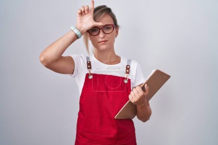 Photo for Young blonde woman wearing waiter uniform holding clipboard making fun of people with fingers on forehead doing loser gesture mocking and insulting. - Royalty Free Image