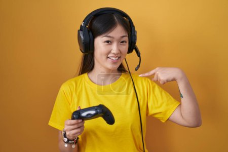 Foto de Chino joven mujer jugando videojuego celebración controlador buscando confiado con sonrisa en la cara, apuntando uno mismo con los dedos orgullosos y felices. - Imagen libre de derechos