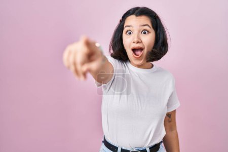 Photo for Young hispanic woman wearing casual white t shirt over pink background pointing with finger surprised ahead, open mouth amazed expression, something on the front - Royalty Free Image