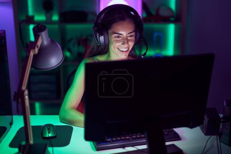 Foto de Young beautiful hispanic woman streamer playing video game using computer at gaming room - Imagen libre de derechos