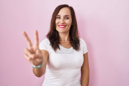 Téléchargez les photos : Femme brune du moyen âge debout sur fond rose souriant en regardant la caméra montrant des doigts faisant signe de victoire. numéro deux. - en image libre de droit