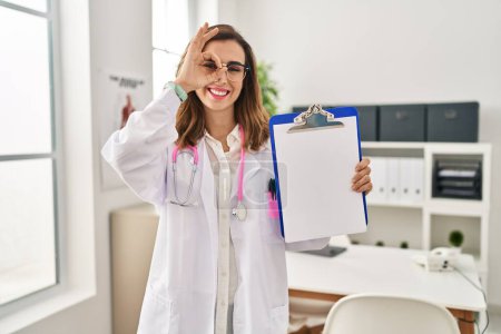 Photo for Young doctor woman holding clipboard smiling happy doing ok sign with hand on eye looking through fingers - Royalty Free Image