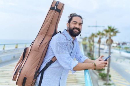Téléchargez les photos : Jeune musicien hispanique utilisant un smartphone tenant un étui de guitare au bord de la mer - en image libre de droit