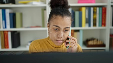 Téléchargez les photos : Jeune étudiante afro-américaine utilisant un ordinateur parlant au téléphone à la bibliothèque - en image libre de droit