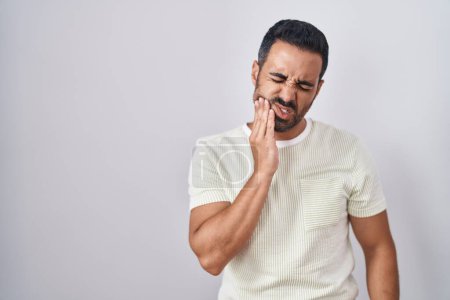 Foto de Hispanic man with beard standing over isolated background touching mouth with hand with painful expression because of toothache or dental illness on teeth. dentist - Imagen libre de derechos