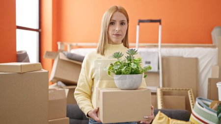 Photo for Young blonde woman holding package and plant with relaxed expression at new home - Royalty Free Image