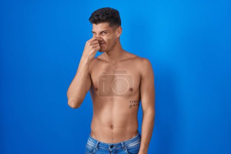 Téléchargez les photos : Jeune homme hispanique debout torse nu sur fond bleu sentant quelque chose de puant et dégoûtant, odeur intolérable, retenant la respiration avec les doigts sur le nez. mauvaise odeur - en image libre de droit