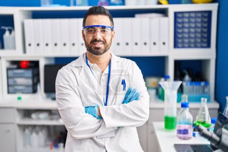 Téléchargez les photos : Jeune homme hispanique scientifique souriant confiant avec les bras croisés geste au laboratoire - en image libre de droit