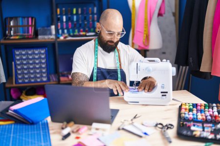 Téléchargez les photos : Jeune tailleur chauve utilisant une machine à coudre et un ordinateur portable à l'usine de vêtements - en image libre de droit